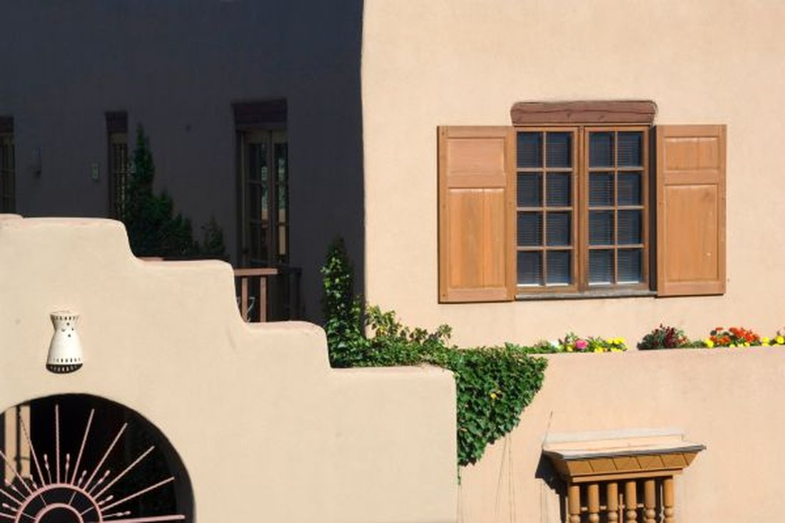 Beige stucco wall with an arched opening, wooden shutters and climbing ivy in warm sunlight
