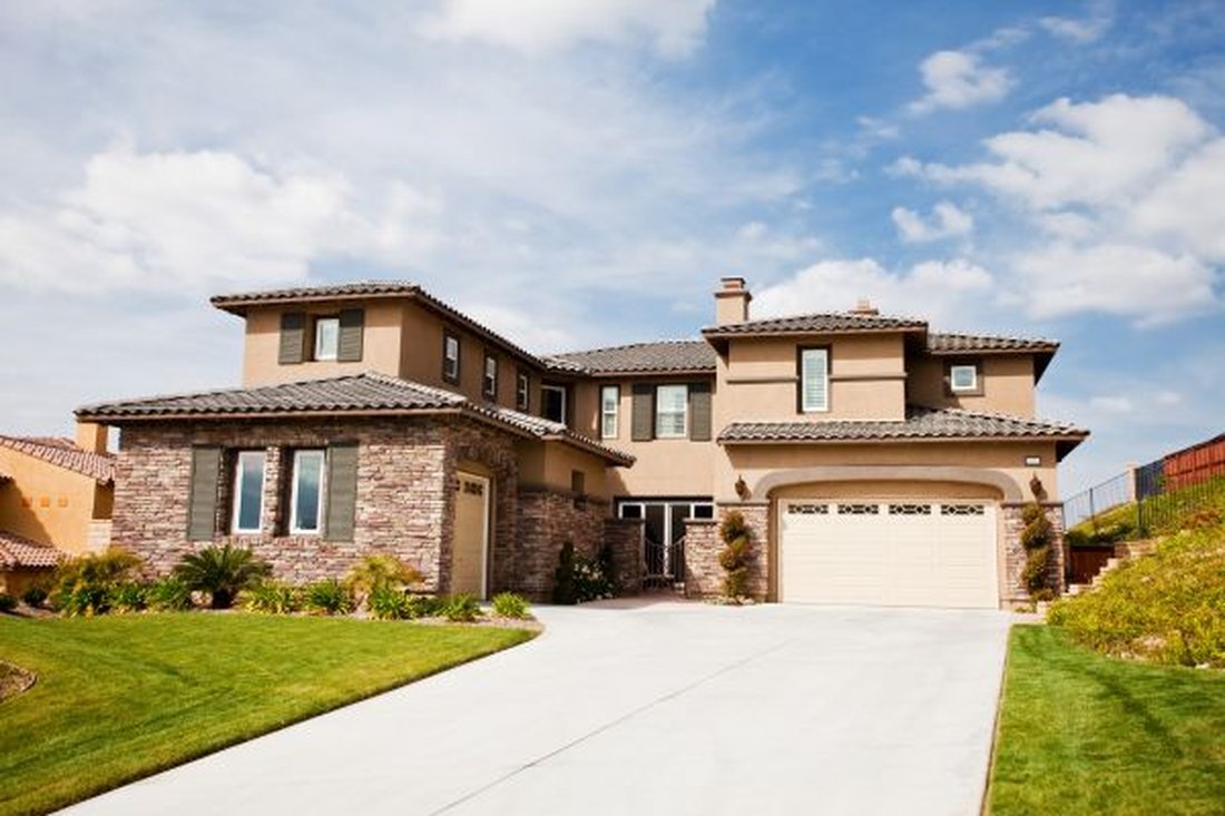 Two-story tan stucco home with stone veneer, dark shutters and a tile roof on a sunny day